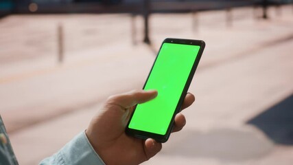 Sticker - Close Up of a Young Man Using Smartphone with Green Screen Chroma Key Mock Up Display in Vertical Position on a Street During a Sunny Day. He's Tapping and Swiping the Screen.