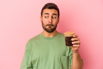 Wall Mural - Young caucasian man holding a coffee bottle isolated on pink background confused, feels doubtful and unsure.