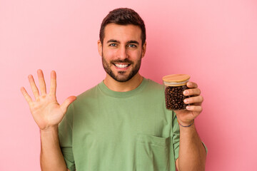 Wall Mural - Young caucasian man holding a coffee bottle isolated on pink background smiling cheerful showing number five with fingers.