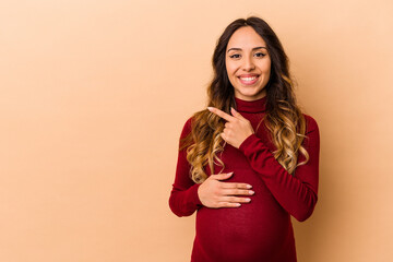 Wall Mural - Young mexican pregnant woman isolated on beige background smiling and pointing aside, showing something at blank space.