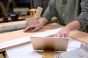 Close-up photo of male carpenter hands using digital tablet during work, work on project of making handmade wooden furniture