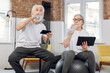 © sofiko14 - Retired man and woman refreshing with water after domestic workout. Senior couple in sport clothes using smartphone and tablet during relaxation.