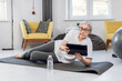 © sofiko14 - Pretty senior woman in sport clothes surfing internet on digital tablet while lying on yoga mat. Active female searching for online tutorial while training at home.