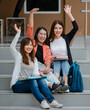 © Bangkok Click Studio - Group of three attractive asian college students sitting down on staircases in university campus waving hello to their friends outdoor. Concept for education, friendship and college students life