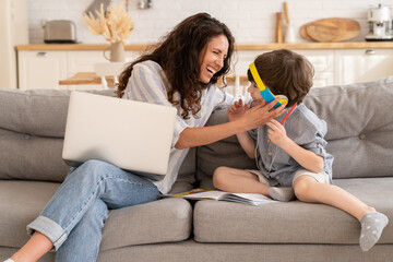 Wall Mural - Happy mom and son laugh sit together on sofa: mother work on laptop and kid in headphones listen to distant school audio lesson, do homework at home at covid-19 quarantine. Family lockdown lifestyle