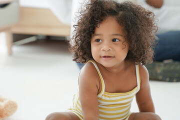 Portrait of happy African little kid girl with curly hair smiling and sitting on the floor at bedroom at home