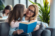 © nenetus - Mature beautiful woman reading a book while her daughter hugging in living room at home.