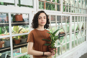 Wall Mural - Portrait of young florist with potted green plant in her hands looking at camera while working in botany garden