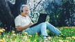 © iana_production - An elderly gray-haired man is working at a laptop, sitting on the grass among yellow flowers in a city park under a tree on a sunny summer day.