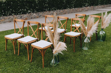 A Row Of Wooden Chairs With Reed Decorations With A Vase Stands On The Green Grass In The Garden, Forest. Wedding Photography.