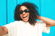 © halayalex - Beautiful black woman with afro curls hairstyle.Smiling hipster model in white t-shirt.Sexy carefree female posing in the street near blue wall in sunglasses.Cheerful and happy.Taking Pov selfie photo