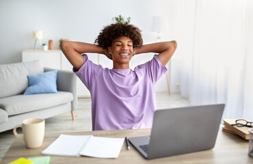 Wall Mural - Cheerful black teen guy holding hands behind head, relaxing in front of laptop at home