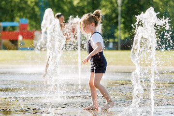 Wall Mural - Cute cheerful girl playing in fountain. Kid in denim overall having fun in summer park