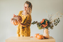 Pretty Blondie Woman In Yellow Sweater Holding Large Orange Pumpkin In Hands While Drawing With Red Marker To Make Traditional Halloween Decor In Light Room At Grey Wall Solid Background