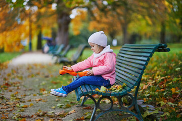 Wall Mural - Adorable little girl sitting on the bench with lunchbox and having picnic on a fall day