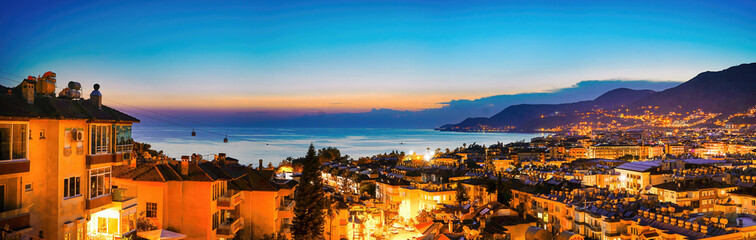 Wall Mural - Wide-format image of Turkish resort of Alanya - beautiful evening sky with reflection in water of bay, silhouette of mountains with scattering of lights and bright evening city, aerial photo.