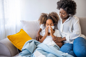 Wall Mural - Mother checking on sick daughter laying in bed. Little girl with cold and blowing her nose. Woman at home blowing the nose of her little girl. Adorable girl blowing nose into tissue paper