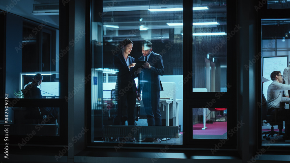 Shot from Outside: Businesswoman and Businessman in a Suit Working on a Tablet Computer in the Office. Manager Standing by the Window. Partners Chat about Financial and Business Development. - obrazy, fototapety, plakaty 