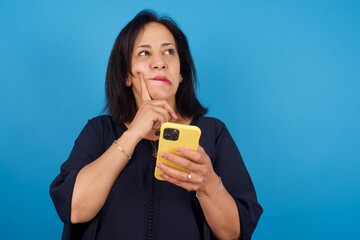 Image of a thinking dreaming middle aged Arab woman standing against blue background using mobile phone and holding hand on face. Taking decisions and social media concept.