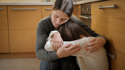 Young mother comforting and calming her daughter crying on floor at kitchen. Concept of domestic violence and family aggression and teenager depression.