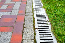 Grating Of The Drainage System For Drainage Of Rainwater In The Park At The Edge Of The Sidewalk From A Stone Slab With A Green Lawn, Landscaping A City Garden Close-up, Nobody.
