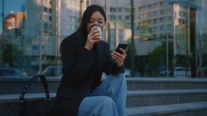 Brunette attractive young modern woman sitting on stairs outside near glass building, browsing smartphone social media and enjoying coffee break.