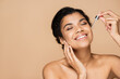 happy african american woman with bare shoulders applying serum isolated on beige