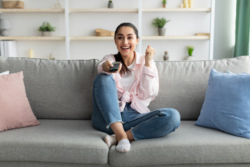 Wall Mural - Excited woman watching tv shaking clenched fist holding remote control