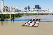 A Tugboat Pushing Barges Full Of Sand And Gravel Down The Ohio River Under A Bridge In Downtown Louisville, Kentucky.
