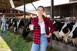 Portrait of young cheerful woman farmer with glass of fresh milk posing at dairy farm
