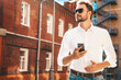 © halayalex - Handsome smiling stylish hipster lambersexual model.Modern man dressed in white shirt. Fashion male posing on the street background in sunglasses. Outdoors at sunset. Using smartphone apps