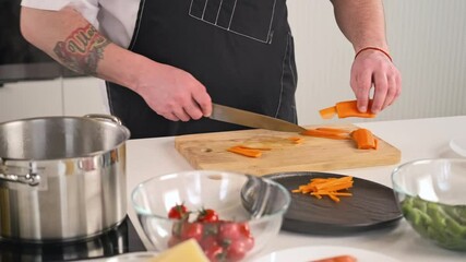 Wall Mural - A male chef is a professional in an apron preparing a salad of vegetables in the kitchen of the restaurant. The cook cuts the raw carrot with a knife on a cutting board. Close-up.