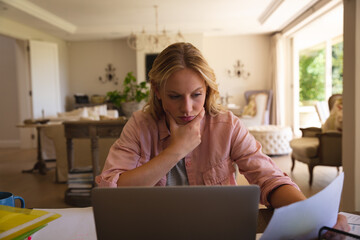 Wall Mural - Caucasian woman sitting at table working in living room using laptop and reading paperwork