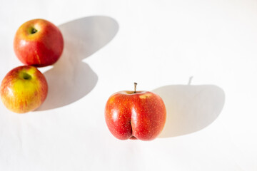 Bright ripe red apple of an unusual shape in the shape of an ass in the light of the sun on a white background with two usual apples behind. Dissimilarity, uniqueness concepts. Top view