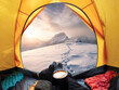Man holding coffee cup and enjoying view of sunrise on snowy mountain inside yellow tent