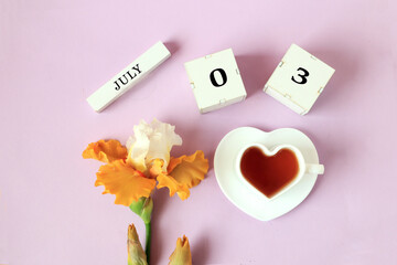 Wall Mural - Calendar for July 3 : the name of the month of July in English, cubes with the numbers 0 and 3, a cup of tea in the shape of a heart, yellow iris on a pastel background, top view