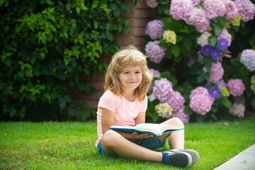 Kid boy reading a book lying on grass. Cute little child in casual clothes reading a book and smiling while lying on grass in park. Learning concept.