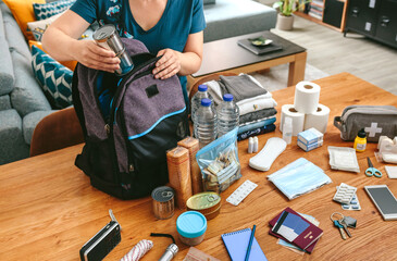 unrecognizable woman putting cans of food to prepare emergency backpack in living room