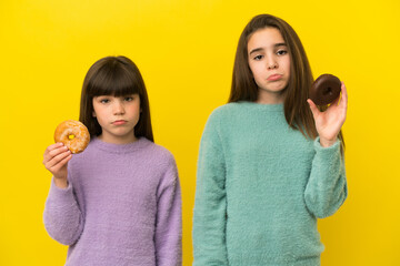 Little sisters isolated on yellow background holding donuts with sad expression