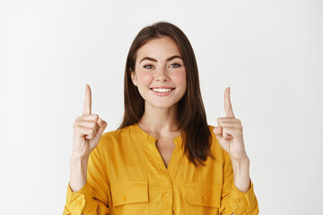 Close-up of smiling young woman showing logo, pointing fingers up and looking confident at camera, standing over white background