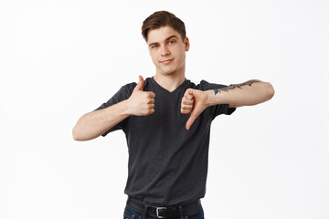 Wall Mural - Judgement. Young guy, college student show thumbs up thumb down, average rate, making decision, like or dislike, standing over white background