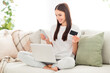 © deagreez - Photo of cheerful lady hold computer debit card texting phone nfc pay wear white t-shirt sit couch in room indoors
