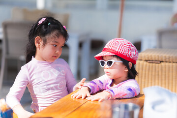 Adorable two Asian girls stand and talk. Child wear sunglasses and a hat. Children go to the sea. Female wear swimsuits. Kid sticks to a table in a restaurant. Baby aged 4-5 years.