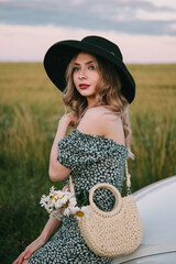 Wall Mural - Beautiful young woman in dress and hat stands.  near a white car.