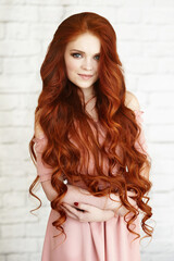 Beautiful unusual young woman in pink dress with long curly red hair on background of white brick. High quality photo