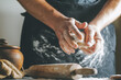 Male hands knead dough with flour next to clay pot and oil bottle and rolling pin on dark table, while cooking