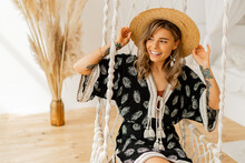 Cozy home atmosphere.  Lovely stylish blond woman in boho dress posing in stylish bedroom. Ladysitting on hanging swing . .