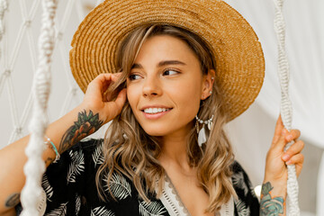 Wall Mural - Smilling young woman in boho dress posing in stylish bedroom.  Sraw hat, boho feather drop earrings . Lady sitting on hanging swing.