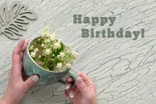 Text Happy Birthday. Old Porcelain Cup In Hands With Pale Yellow Wild Flowers. Aged Crackled Paint, Flat Lay On Textured Light Mint Green Background. Mature Female Hands Hold The Cup. Floral Wood