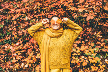 Playful Young Woman In Bright Clothes With Long Scarf Hides Eyes Behind Dry Orange Leaves Near Large Red Bush In Picturesque Autumn Forest